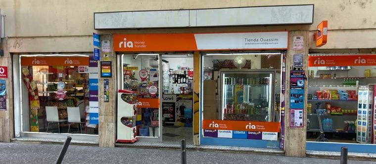Fachada de Tienda Ouassim en Carrer del Progrés, 5: el rótulo naranja de Ria Money Transfer, el letrero blanco con el nombre de la tienda y los escaparates con las marcas de recargas